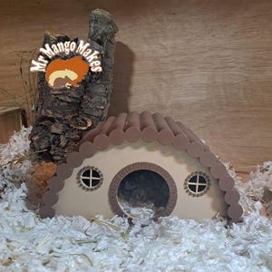 May include: A brown and beige hamster house with a rounded roof and two small windows. The house has a large opening in the front for the hamster to enter and exit. The house is sitting on a bed of white bedding.