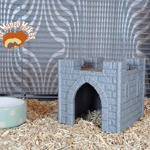 May include: A grey stone castle-shaped hideaway for small pets, with a rounded archway entrance. The hideaway is sitting on a bed of brown straw-like bedding.