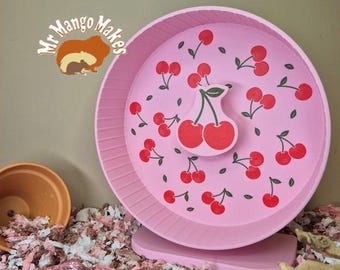 May include: A pink hamster wheel with a cherry design. The wheel has a pink base and is decorated with red cherries and green leaves. The wheel is sitting on a bed of shredded paper bedding.