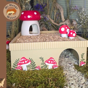 May include: A beige rectangular small animal hideout with a ribbed top edge and a large arched entrance. The exterior features printed red and white mushroom and green foliage graphics. A ceramic red mushroom house sits on top.