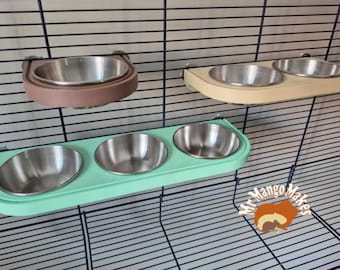 May include: A collection of pet food bowls in various colors, designed to attach to a cage. The bowls are made of stainless steel and are set in a brown, cream, and mint green holder. The cage is made of black metal.