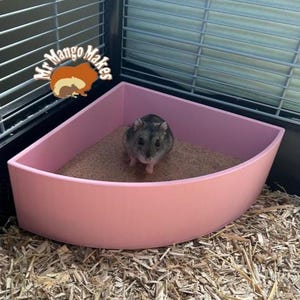 May include: A small grey hamster in a pink corner litter tray filled with sand. The tray is inside a wire cage. The cage sits on wood shavings. The logo "Mr Mango Makes" is in the top left corner.