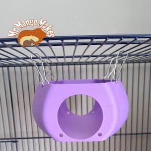 May include: A purple hanging cage accessory for small pets. The accessory is shaped like a rounded square with a hole in the centre. It is suspended from the top of a cage by two metal clips.