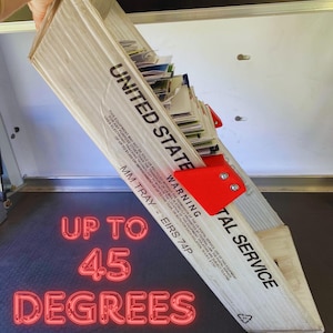 May include: A cardboard United States Postal Service mail tray, tilted at a 45-degree angle, filled with mail. The tray has red brackets and the text "UNITED STATE" and "POSTAL SERVICE". The text "UP TO 45 DEGREES" is also visible.