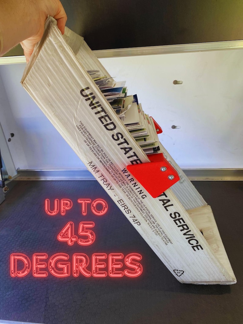 May include: A cardboard United States Postal Service mail tray, tilted at a 45-degree angle, filled with mail. The tray has red brackets and the text "UNITED STATE" and "POSTAL SERVICE". The text "UP TO 45 DEGREES" is also visible.