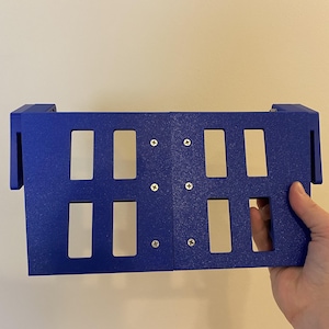 May include: A blue rectangular object with eight rectangular cutouts and four screws. The object is held by a hand. The object has two side extensions.