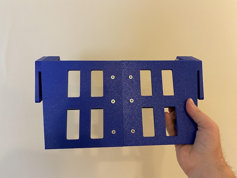 May include: A blue rectangular object with eight rectangular cutouts and four screws. The object is held by a hand. The object has two side extensions.