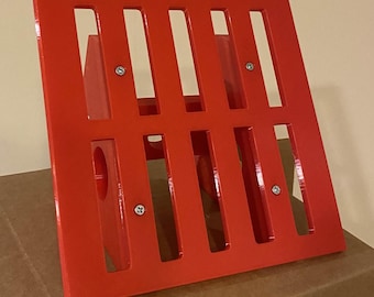 May include: A red metal grate with a rectangular shape and multiple vertical slots. The grate is attached to a red metal frame with four screws visible.