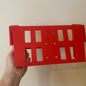 May include: A bright red rectangular object with multiple rectangular cutouts. The object has a grid-like appearance and is held in front of a neutral-colored wall. The object is made of a solid material and has a textured surface.