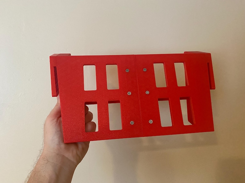 May include: A bright red rectangular object with multiple rectangular cutouts. The object has a grid-like appearance and is held in front of a neutral-colored wall. The object is made of a solid material and has a textured surface.