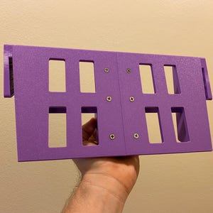 May include: A purple rectangular object with multiple rectangular cutouts, held in a hand. The object is constructed from two connected panels, secured with screws. The object has a textured surface and is designed with protruding edges.