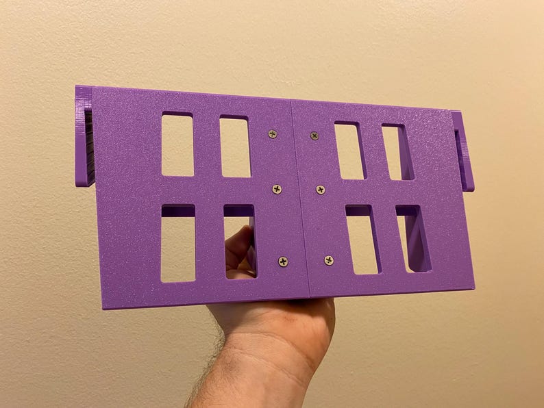 May include: A purple rectangular object with multiple rectangular cutouts, held in a hand. The object is constructed from two connected panels, secured with screws. The object has a textured surface and is designed with protruding edges.