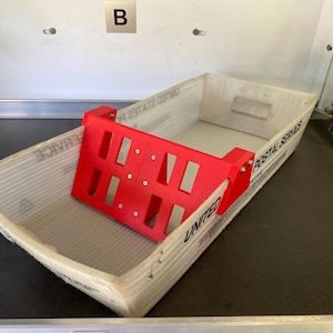 May include: A red 3D-printed mail sorter sits inside a corrugated cardboard box. The sorter has a rectangular shape with multiple cut-out sections. The box is labeled "UNITED POSTAL SERVICE".
