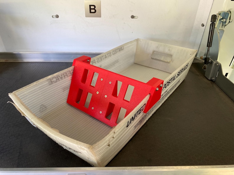 May include: A red 3D-printed mail sorter sits inside a corrugated cardboard box. The sorter has a rectangular shape with multiple cut-out sections. The box is labeled "UNITED POSTAL SERVICE".
