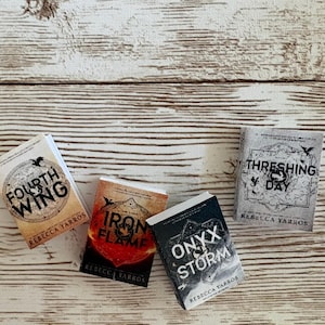 May include: Four miniature books by Rebecca Yarros titled Fourth Wing, Iron Flame, Onyx Storm, and Threshing Day. Each book features a distinct cover design with black text and graphic illustrations, arranged in a cluster on a light wood surface.