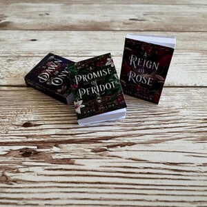 May include: Three miniature books with titles: "A Dawn of Onyx", "A Promise of Peridot", and "A Reign of Rose". The books have colorful covers with floral and fantasy-themed designs. They are arranged on a light brown wooden surface.
