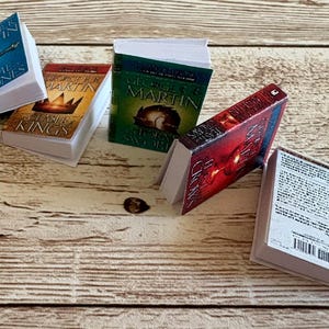 K&ouml;nnte beinhalten: Miniatur-Buchset mit Titeln wie "A Game of Thrones" und "A Clash of Kings". Die B&uuml;cher haben wei&szlig;e Seiten und sind auf einer Holzoberfl&auml;che angeordnet. Die Einb&auml;nde sind in Blau, Rot, Gr&uuml;n und Gold gehalten.