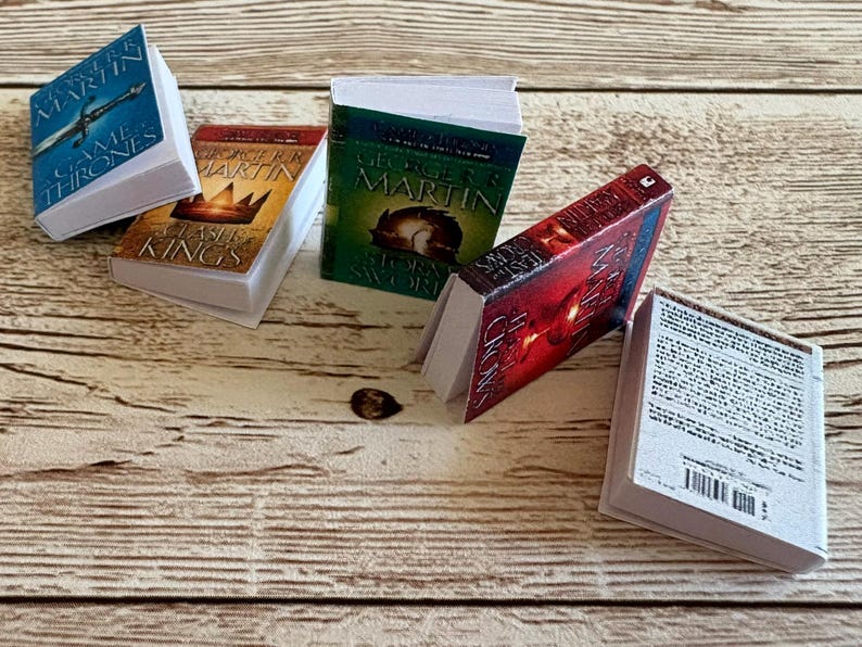 K&ouml;nnte beinhalten: Miniatur-Buchset mit Titeln wie "A Game of Thrones" und "A Clash of Kings". Die B&uuml;cher haben wei&szlig;e Seiten und sind auf einer Holzoberfl&auml;che angeordnet. Die Einb&auml;nde sind in Blau, Rot, Gr&uuml;n und Gold gehalten.