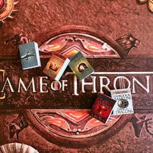 K&ouml;nnte beinhalten: Nahaufnahme eines Brettspiels mit dem Schriftzug "Game of Thrones" in erhabenen Buchstaben. Mehrere Miniaturb&uuml;cher, darunter "Game of Thrones" und "A Clash of Kings", liegen auf dem Spielbrett und repr&auml;sentieren die Fantasy-Buchreihe.