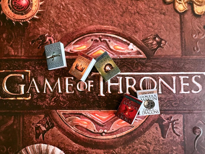 K&ouml;nnte beinhalten: Nahaufnahme eines Brettspiels mit dem Schriftzug "Game of Thrones" in erhabenen Buchstaben. Mehrere Miniaturb&uuml;cher, darunter "Game of Thrones" und "A Clash of Kings", liegen auf dem Spielbrett und repr&auml;sentieren die Fantasy-Buchreihe.