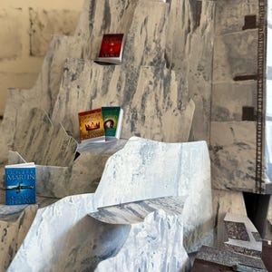 K&ouml;nnte beinhalten: Ein Miniatur-Diorama eines Gebirges mit mehreren kleinen B&uuml;chern, die auf den Gipfeln ausgestellt sind. Die B&uuml;cher haben farbenfrohe Einb&auml;nde und Titel wie "Game of Thrones" und "A Dance with Dragons". Das Diorama besteht aus grauem und wei&szlig;em Papier.