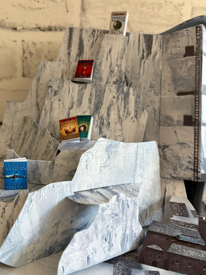K&ouml;nnte beinhalten: Ein Miniatur-Diorama eines Gebirges mit mehreren kleinen B&uuml;chern, die auf den Gipfeln ausgestellt sind. Die B&uuml;cher haben farbenfrohe Einb&auml;nde und Titel wie "Game of Thrones" und "A Dance with Dragons". Das Diorama besteht aus grauem und wei&szlig;em Papier.