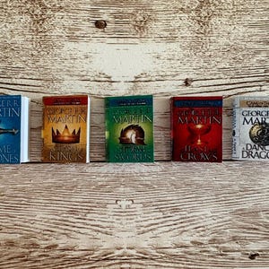 K&ouml;nnte beinhalten: F&uuml;nf Miniaturb&uuml;cher aus der "A Song of Ice and Fire"-Reihe von George R.R. Martin. Die B&uuml;cher haben farbenfrohe Einb&auml;nde mit Titeln wie "A Game of Thrones" und "A Clash of Kings". Sie sind auf einer hellbraunen Holzoberfl&auml;che angeordnet.