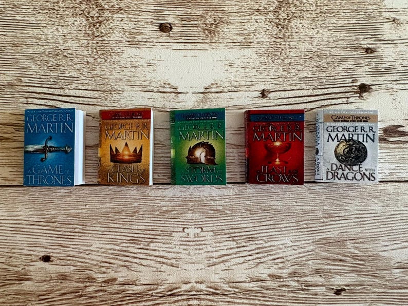K&ouml;nnte beinhalten: F&uuml;nf Miniaturb&uuml;cher aus der "A Song of Ice and Fire"-Reihe von George R.R. Martin. Die B&uuml;cher haben farbenfrohe Einb&auml;nde mit Titeln wie "A Game of Thrones" und "A Clash of Kings". Sie sind auf einer hellbraunen Holzoberfl&auml;che angeordnet.