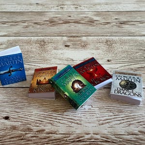 K&ouml;nnte beinhalten: Miniaturb&uuml;cher aus der "A Song of Ice and Fire"-Reihe von George R.R. Martin. Die B&uuml;cher haben farbenfrohe Einb&auml;nde mit Titeln wie "Game of Thrones" und "A Storm of Swords". Sie sind auf einer hellen Holzmaserung angeordnet.