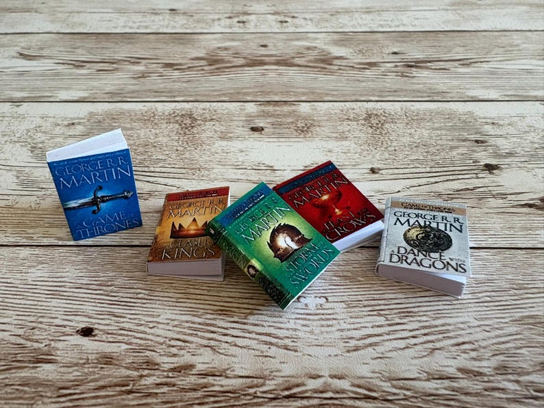K&ouml;nnte beinhalten: Miniaturb&uuml;cher aus der "A Song of Ice and Fire"-Reihe von George R.R. Martin. Die B&uuml;cher haben farbenfrohe Einb&auml;nde mit Titeln wie "Game of Thrones" und "A Storm of Swords". Sie sind auf einer hellen Holzmaserung angeordnet.