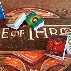K&ouml;nnte beinhalten: Miniaturb&uuml;cher mit farbenfrohen Einb&auml;nden, darunter Titel wie "Game of Thrones" und "A Feast for Crows" von George R.R. Martin. Die B&uuml;cher liegen auf einer braunen Oberfl&auml;che mit dem Schriftzug "Game of Thrones".