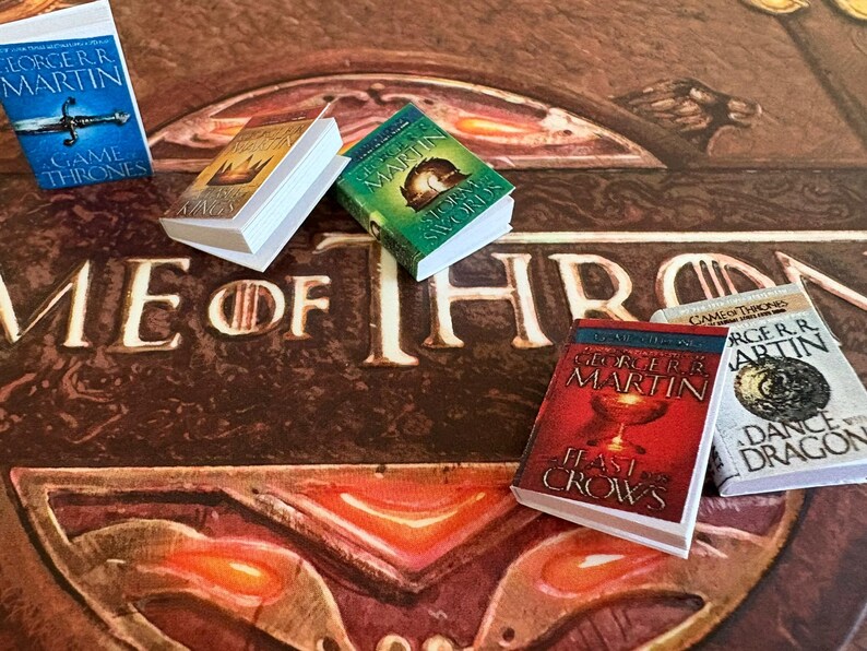 K&ouml;nnte beinhalten: Miniaturb&uuml;cher mit farbenfrohen Einb&auml;nden, darunter Titel wie "Game of Thrones" und "A Feast for Crows" von George R.R. Martin. Die B&uuml;cher liegen auf einer braunen Oberfl&auml;che mit dem Schriftzug "Game of Thrones".