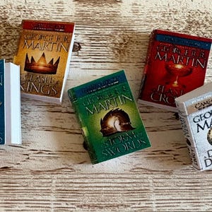 K&ouml;nnte beinhalten: Miniatur-Buchset mit Titeln von George R.R. Martin, darunter "Game of Thrones", "Clash of Kings", "Storm of Swords", "Feast for Crows" und "Dance with Dragons". Die B&uuml;cher haben farbenfrohe Einb&auml;nde und wei&szlig;e Seiten.