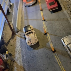 以下が含まれることがあります： 灰色の舗装道路にミニチュアの車が並んだシーン。ナンバー53のベージュ色のフォルクスワーゲンビートル、赤いスポーツカー、オレンジ色のAlliedトラックが見えます。オレンジ色のコーンが道路に並んでいます。
