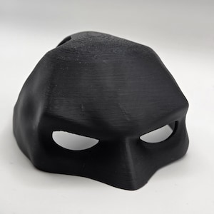 Puede incluir: Una máscara negra moldeada con aberturas para los ojos. La máscara tiene una superficie texturizada y una parte superior redondeada. Probablemente para disfraces o cosplay.