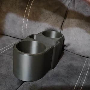 May include: A dark gray, 3D-printed cup holder with two circular openings and a smaller rectangular compartment. The cup holder is designed to hold two cups or bottles and is made of a smooth, solid material.