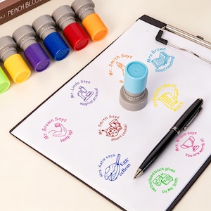 以下が含まれることがあります： YOU SAY PEACH BLOSSOMと書かれた茶色の箱に入った、カラフルなセルフインクスタンプのセット。スタンプのキャップはピンク、緑、黄色、紫、青、赤、オレンジなど様々な色があります。クリップボードにはスタンプされた文字やアイコンが並んでいます。