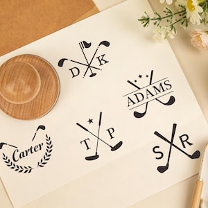 May include: A round wooden rubber stamp sits on a white sheet of paper displaying five black ink impressions. The designs feature crossed golf clubs paired with the names Carter and Adams, or the initials D K, T P, and S R, accented by stars.