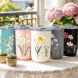 Könnte beinhalten: Eine Sammlung isolierter Becher in verschiedenen Farben, jeweils mit floralen Designs und personalisierten Namen verziert. Die Becher haben einen silbernen Rand, einen grauen Deckel und einen transparenten Schiebeverschluss. Die Farben umfassen Türkis, Rosa, Weiß, Grau und Schwarz.