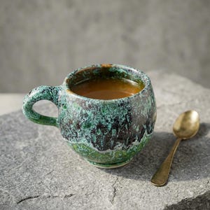 May include: A handmade ceramic mug with a green and black glaze, filled with a warm beverage. A small, gold-colored spoon rests beside the mug on a gray stone surface. The mug has a rounded shape and a sturdy handle.