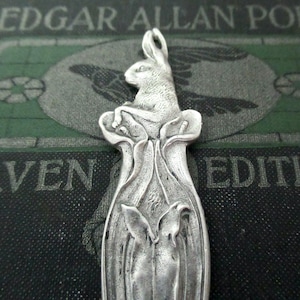 May include: A silver-toned pendant featuring a detailed rabbit perched atop a stylized floral design. The pendant has a vintage aesthetic, with intricate detailing on the rabbit and the surrounding floral elements. The background is a dark green and black book cover.
