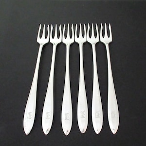 May include: A set of six silver-toned metal cocktail forks arranged in a row. Each fork features a three-pronged head and a smooth, tapered handle. A small, engraved monogram consisting of the letters 'ME' is visible on the lower part of each handle.