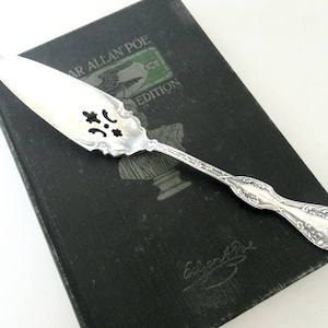 May include: A silver cake server with an ornate handle and a decorative, slotted blade rests on a dark green book. The book has the text "EDGAR ALLAN POE EDITION" and a signature. The server has a floral design.