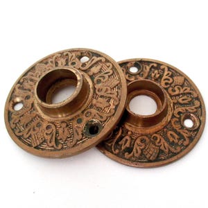 May include: Two circular brass door escutcheons featuring an intricate embossed floral and scrollwork pattern. Each plate has a central opening for a spindle and two small holes for mounting screws, with a weathered, aged metallic finish.