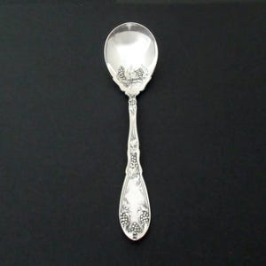May include: A silver spoon with a rounded bowl and an ornate handle. The spoon features a grape cluster design on the bowl and handle. The spoon is likely made of sterling silver and is displayed against a black background.