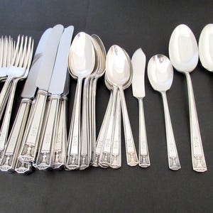 May include: A collection of silver-colored flatware, including forks, knives, spoons, and butter knives, arranged on a dark surface. The utensils have decorative handles with a repeating pattern. The spoons and forks are arranged in a fan shape.