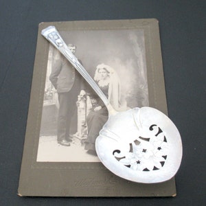 May include: A silver serving spoon with an ornate design and a cutout pattern. The spoon is resting on top of an old photograph of a couple.