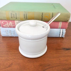 May include: A white ceramic sugar bowl with a lid and a small spoon. The bowl is round with a slightly flared base and a small knob on the lid. The spoon is clear. The bowl is on a wooden surface with books in the background.