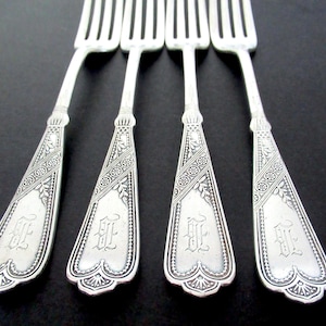 May include: Four silver forks with ornate, engraved handles. The handles feature a decorative floral and geometric pattern, with an initial in the center. The forks are arranged on a dark surface.