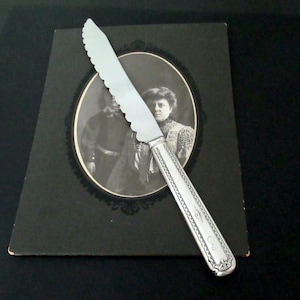 May include: A silver butter knife with a serrated blade rests on a vintage black and white photograph. The knife has a decorative handle with engraved details. The photograph is in a black frame.